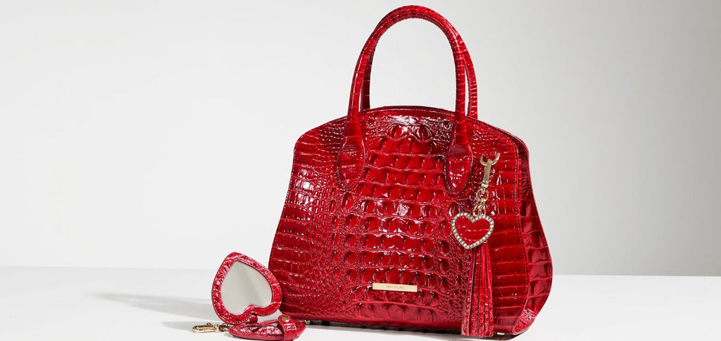Red croc-embossed leather structured satchel handbag with rolled top handles, gold-tone logo bar, heart-shaped charm, and leather tassel, photographed on a white background