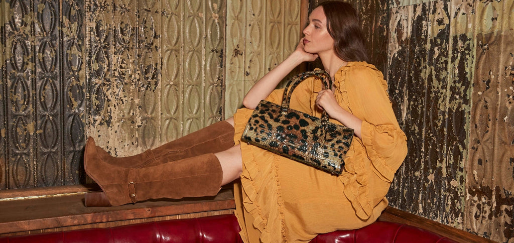 Woman in a mustard ruffled dress reclining on a red leather banquette while holding a Brahmin leopard-print croc-embossed satchel, styled against a vintage textured wall.