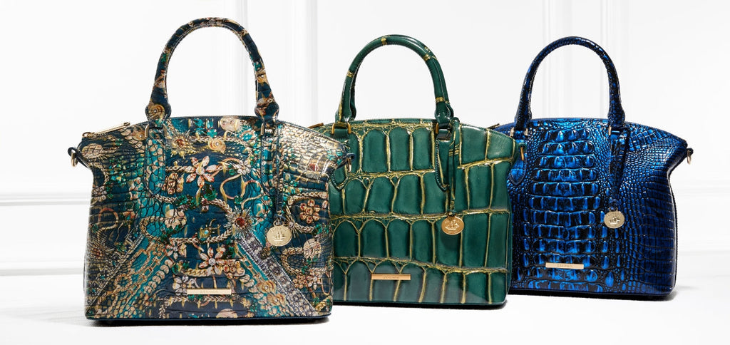 Three Brahmin structured satchel handbags displayed side by side: a floral-inspired teal multi embossed satchel with intricate gold detailing, a deep green croc-embossed satchel with gold accents, and a vibrant sapphire blue croc-embossed satchel.