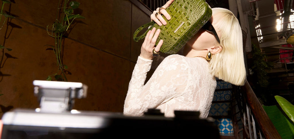 Blonde woman in a sheer lace top holding an olive green crocodile embossed handbag in front of her face in a warm indoor setting