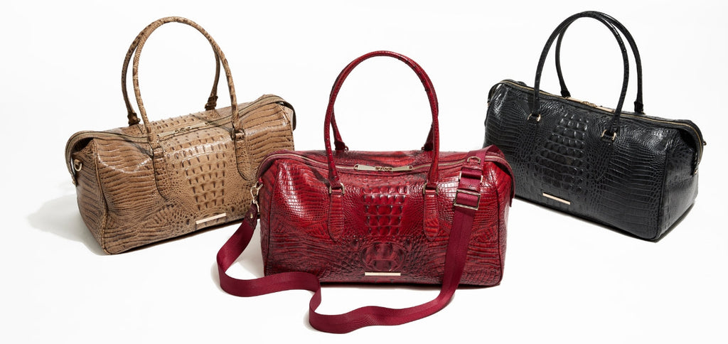 Three Brahmin croc-embossed duffle bags displayed in taupe, deep red, and classic black, each with structured handles and gold hardware, photographed on a clean white background.