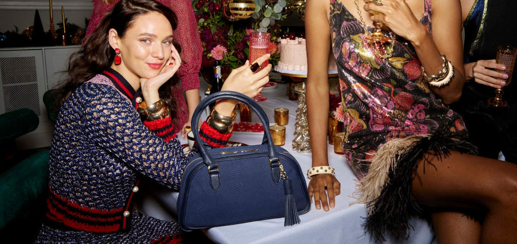 Women gathered at a holiday party wearing festive dresses, with a navy blue textured leather satchel handbag displayed on the table in the foreground.
