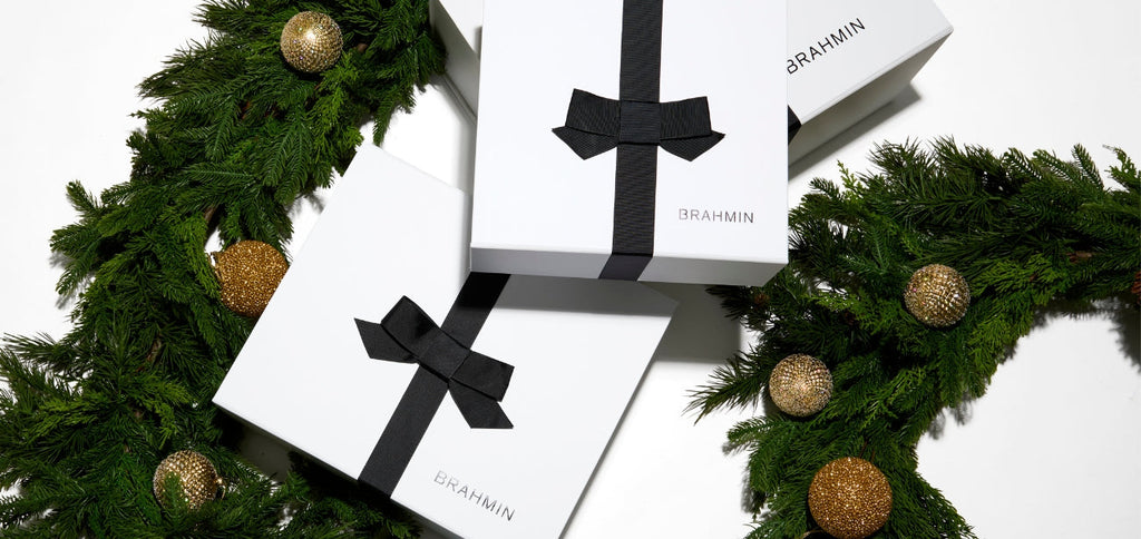 White Brahmin gift boxes tied with black bows arranged among holiday greenery and gold ornaments, creating an elegant festive presentation