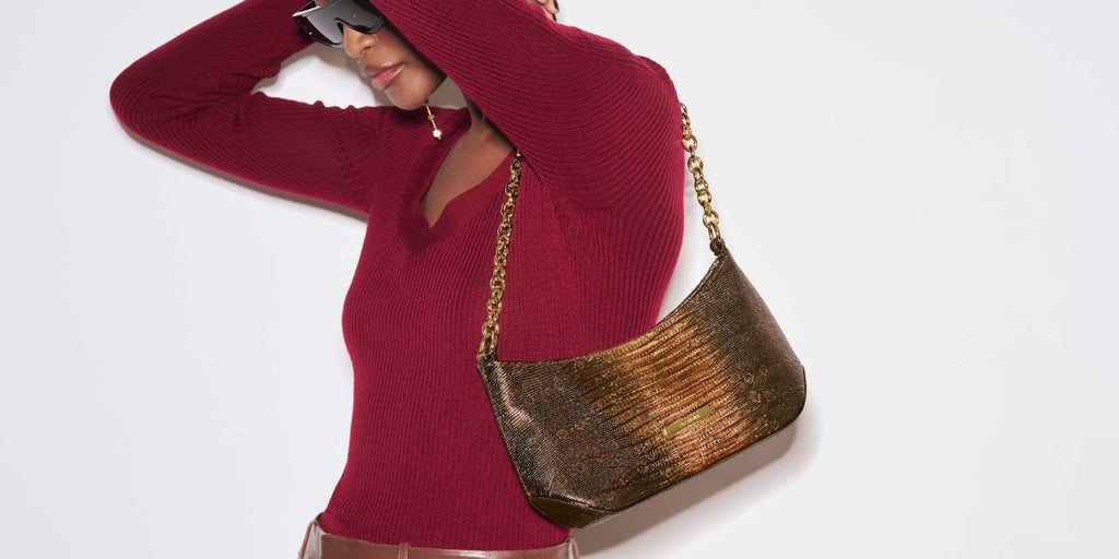 Model in a burgundy sweater and brown leather pants adjusting her hair while wearing a small brown Brahmin shoulder bag with a chain strap. 