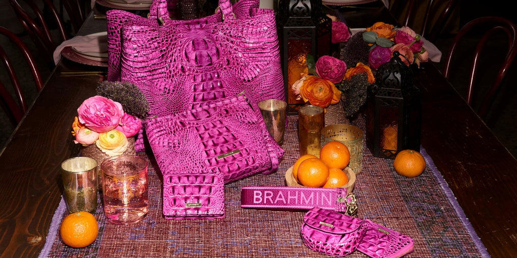 Bright pink Brahmin Melbourne collection displayed on a dining table with a crocodile embossed tote, matching pouch, wallet, strap, small accessories, flowers, candles, and oranges