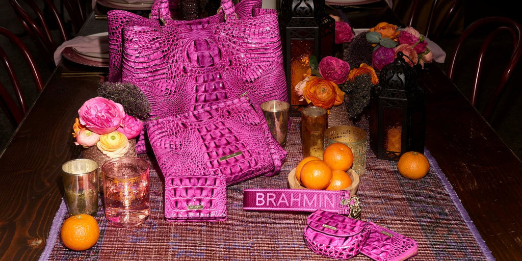 Bright pink Brahmin Melbourne collection displayed on a dining table with a crocodile embossed tote, matching pouch, wallet, strap, small accessories, flowers, candles, and oranges