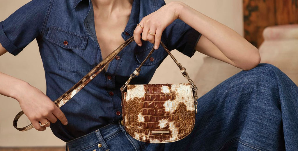 Model in dark denim shirt and jeans holding a Brahmin Western Trend embossed croc leather saddle bag in metallic cognac and cream, styled against a neutral studio background.