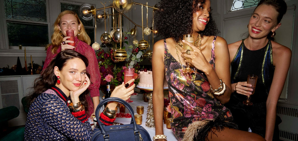 Holiday party with four women in festive outfits, sipping cocktails around a table with a navy croc-embossed satchel.