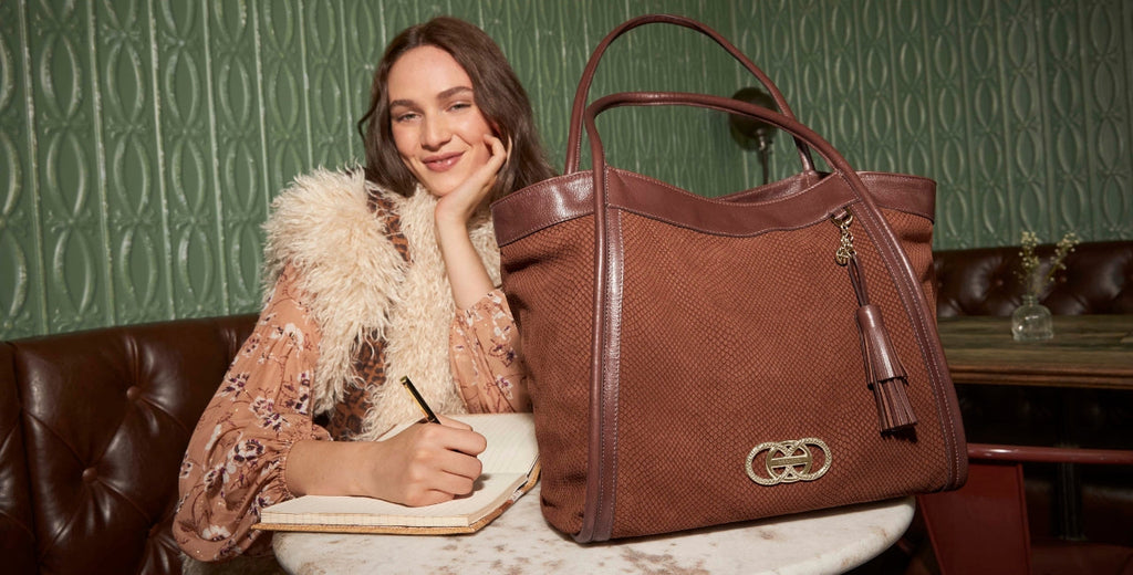 Woman writing at a café beside an oversized brown textured leather tote with tassel and signature hardware.