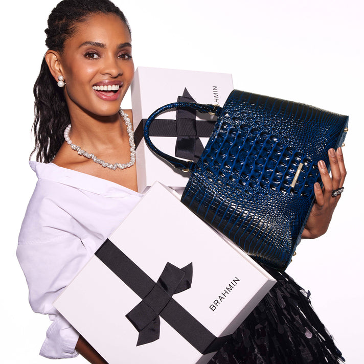 Model holding a deep blue Brahmin structured handbag with glossy embossed texture, double top handles and gold tone hardware beside Brahmin gift boxes.