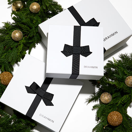 Stack of Brahmin gift boxes wrapped in white paper with black ribbon bows, displayed among decorated Christmas tree branches with gold ornaments for a festive holiday presentation.