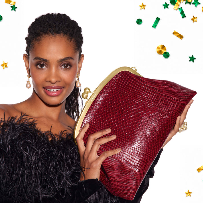 Model holding a large red Brahmin clutch bag with textured finish, oversized gold tone frame closure and festive green and gold confetti.