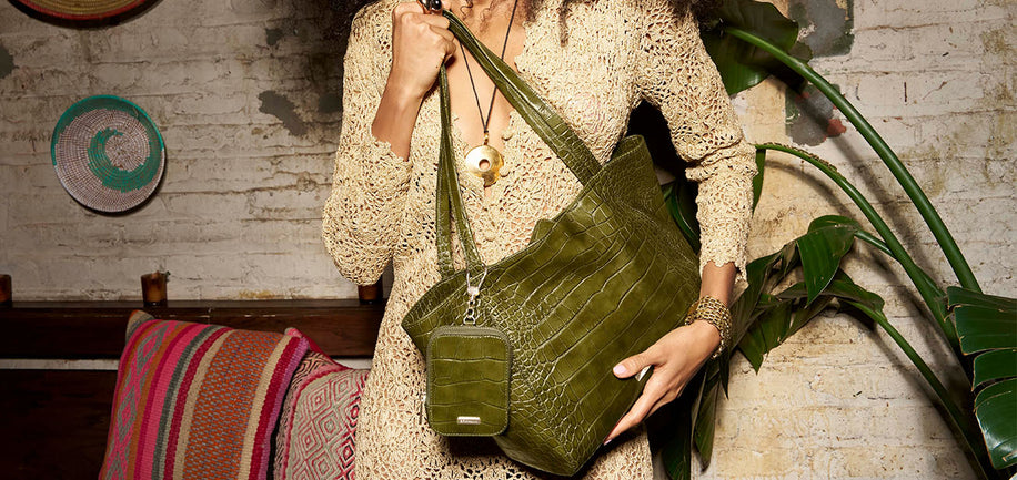 Person holding a green handbag in a casual setting with a plant and colorful pillow in the background.
