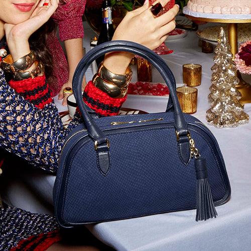 Brahmin Soft & Slouchy handbag in deep navy leather, featuring a rounded relaxed shape, rolled handles, and a detachable tassel charm, styled on a holiday table setting.