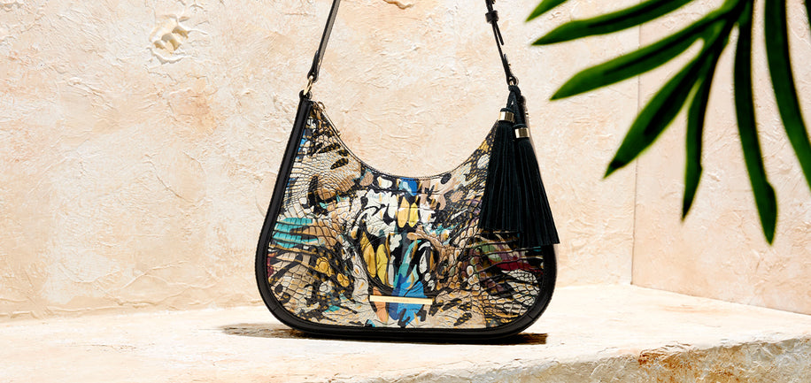A shoulder bag with a black tassel and a colorful abstract graffiti-style print, resting on a stone ledge against a textured beige wall with a green palm leaf in the background.