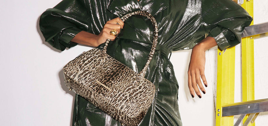 A model in a shiny dark green dress holds a structured leopard-print handbag with a woven handle, standing next to a yellow ladder against a white wall.