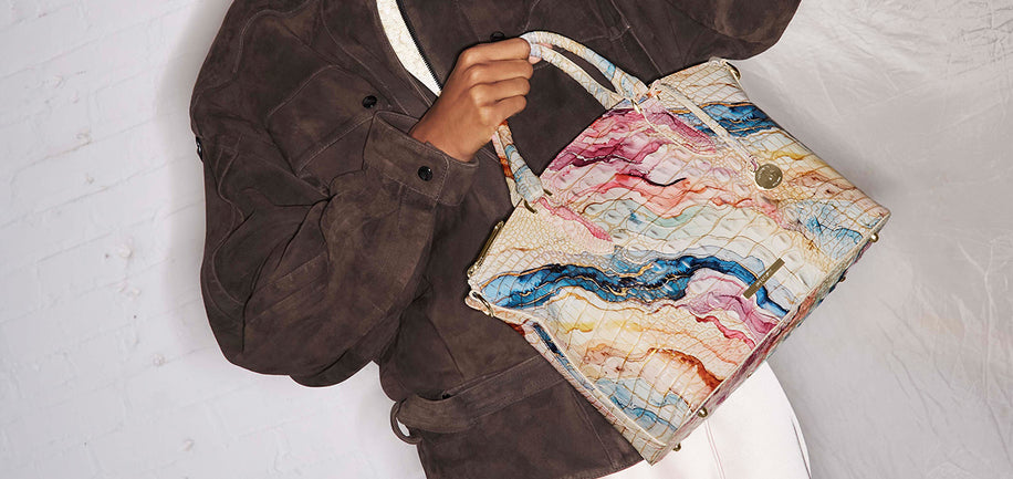 A person wearing a brown suede jacket holds a large handbag with a colorful marble-like pattern in cream, pink, blue, and orange against a white textured background.