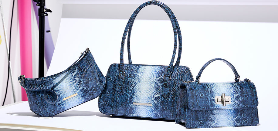 Three blue snakeskin-patterned handbags displayed together on a white surface against a white background, including a large top-handle satchel, a smaller top-handle satchel, and a crescent shoulder bag.