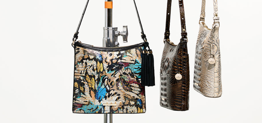 Three Brahmin shoulder bags displayed on a white background, including a multicolor abstract print bag on a stand, a dark brown crocodile-embossed bag, and a beige croc embossed bag.