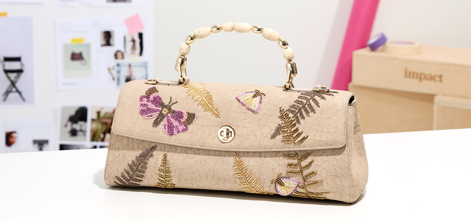 A beige textured handbag with a top handle made of cream-colored beads, decorated with gold and purple butterfly and fern embroidery. The bag features a central gold-tone clasp and is displayed on a white surface with a blurred studio background.