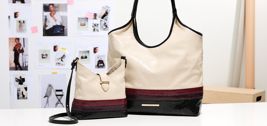 A beige tote bag with black trim and a matching small shoulder bag, both featuring a dark burgundy and black textured bottom panel, displayed on a white table with a fashion board in the background.