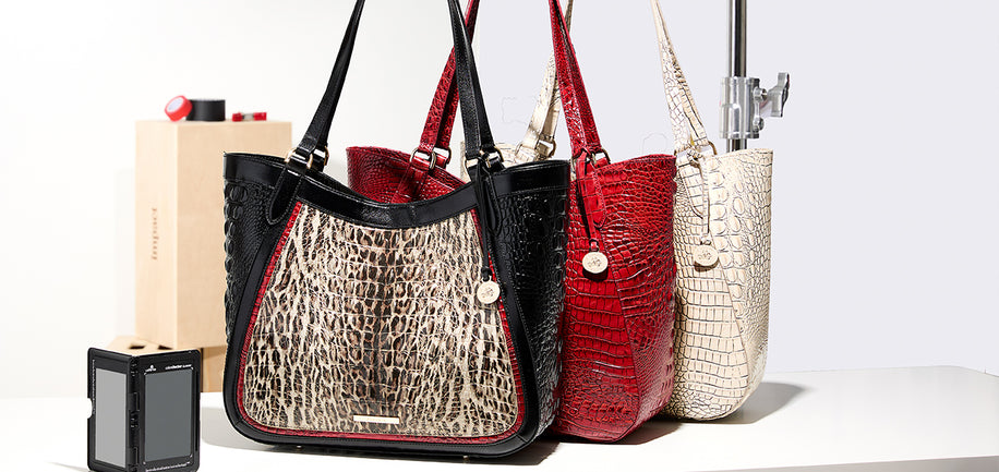 Three handbags lined up on a white surface against a light gray background: a black tote with a beige-and-black crocodile-embossed front panel, a red crocodile-embossed tote, and a cream crocodile-embossed tote. A small camera and a cardboard box are placed beside them as props.