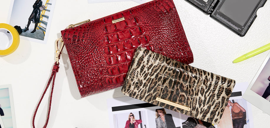 Two designer wallets displayed on a white surface with fashion magazine pages. The top-left clutch is a vibrant red with an embossed crocodile pattern and a wristlet strap.