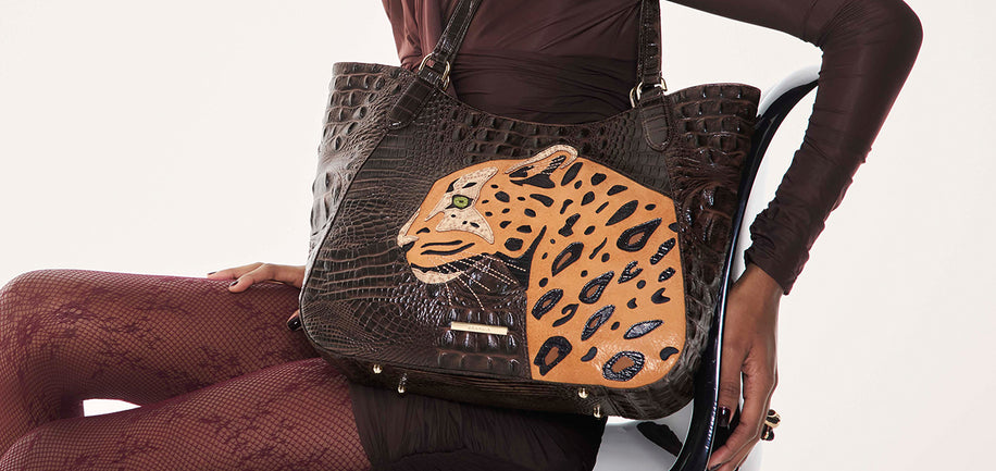 Sheer brown outfit seated holding a Brahmin satchel handbag with embossed pattern and painted leopard face design, structured silhouette, top handles, gold-tone hardware, textured finish, front view.