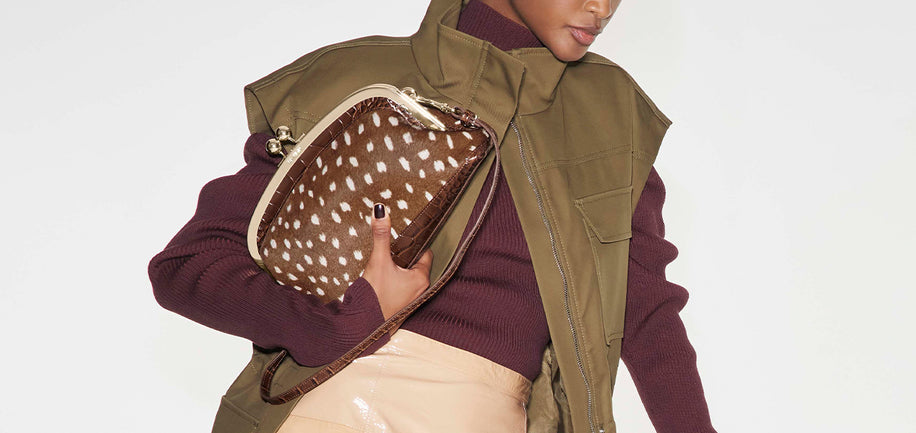 Model holds Brahmin brown patterned clutch with structured silhouette, flap closure, and metallic hardware visible. Upper body shown in burgundy turtleneck and olive vest against a plain background.