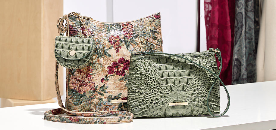 A styled product display featuring two designer handbags: a green crocodile-embossed leather shoulder bag in the foreground and a floral tapestry-patterned hobo bag in the background, set on a white surface with a clothing rack blurred in the background.