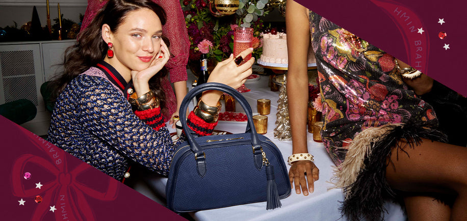 Angie Navy Belcanto suede satchel in rich navy is embossed with a snake print and trimmed with a matching smooth leather for a monochromatic moment sitting on a table full of holiday accessories and model holding a glass looking at the camara