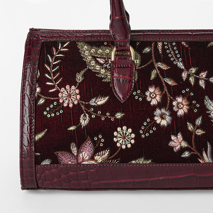 A close-up of a structured handbag featuring deep burgundy intricate floral embroidery in pink, green, and gold, framed by textured crocodile-embossed burgundy leather trim and a matching top handle.