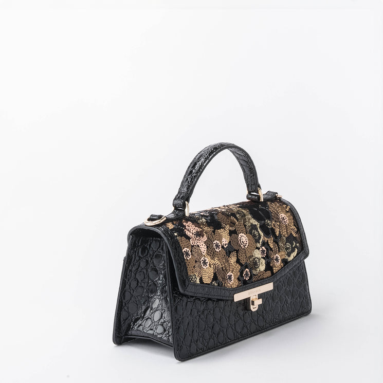 BRAHMIN The Genie mini satchel in black with embossed texture, gold clasp closure, top handle, floral sequin flap, crossbody strap, and structured silhouette shown on a white background.