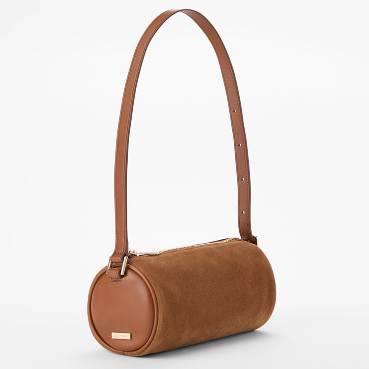 A brown cylindrical shoulder bag with a smooth suede-like finish, a matching adjustable shoulder strap, and a small rectangular metal plaque on the front, shown against a plain white background.
