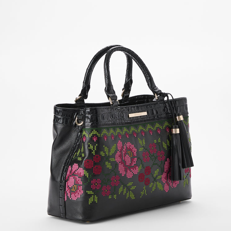 Brahmin black leather satchel with double top handles, floral pink and green cross-stitch embroidery on the front, gold-tone hardware, two black tassels, structured silhouette, and decorative front details.
