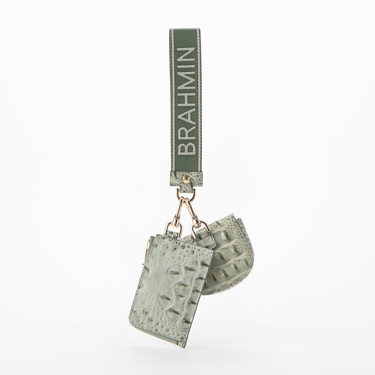 A light sage green leather keychain bag with textured crocodile and alligator patterns, featuring a matching strap with the brand name 'BRAHMIN' in white lettering, suspended from gold-tone hardware.