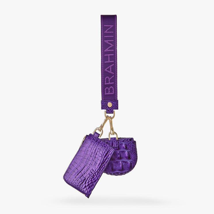 Brahmin purple wristlet strap with gold hardware holds a small textured card case and matching round pouch, both in purple faux leather with embossed pattern, displayed on a white background.