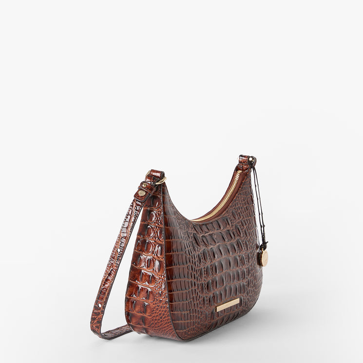 A brown crocodile-embossed shoulder bag with a crescent shape, a long adjustable strap, and a small gold-tone metal plaque on the front, shown against a plain light gray background. Side view.