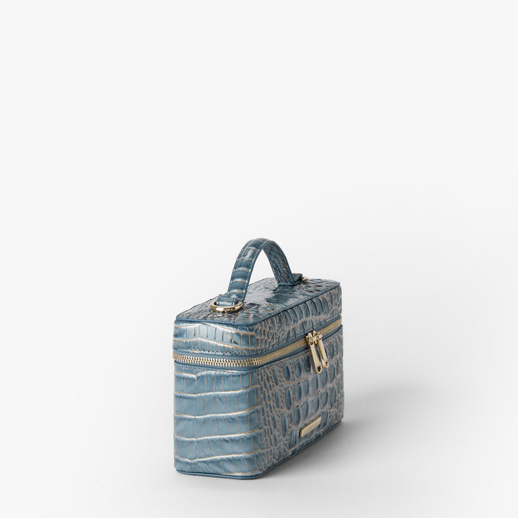 A light blue crocodile-embossed leather cosmetic case with a top handle, gold-tone zipper, and a small gold Brahmin plaque on the front, set against a plain white background. Side view.