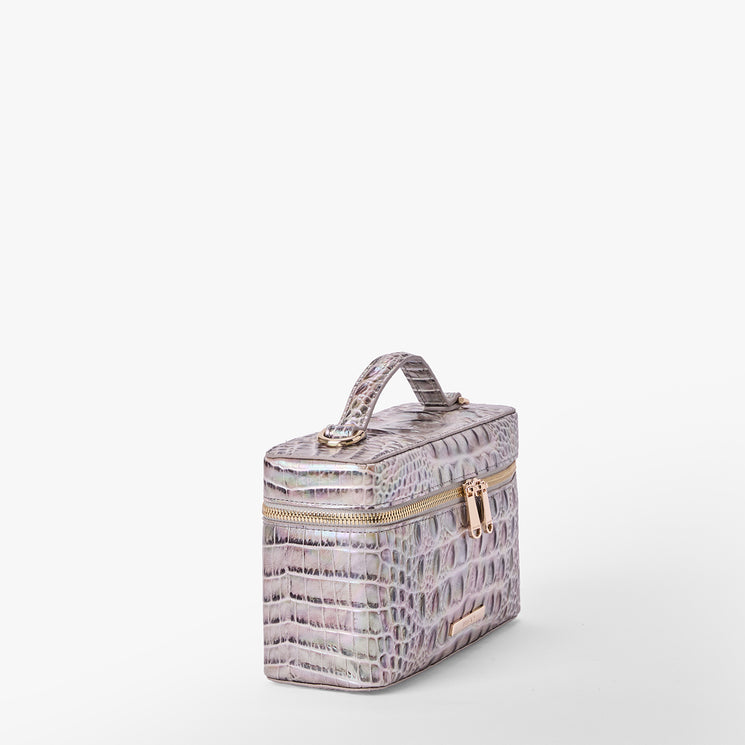 Brahmin cosmetic case in pastel purple and beige with embossed textured pattern, rectangular silhouette, gold-tone zipper, matching top handle. Shown against a plain white background.
