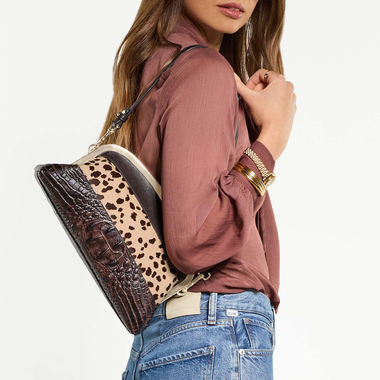 A woman wearing a dusty rose blouse and blue jeans carries a shoulder bag with a dark brown crocodile-embossed front, a beige leopard-print body, and a black strap, against a plain white background.