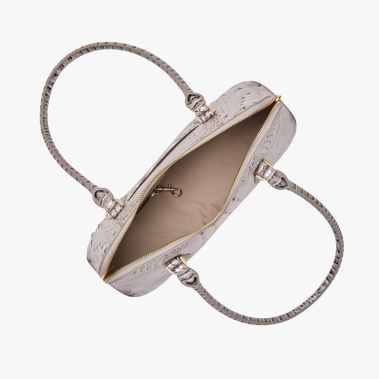 Top view of a Brahmin light gray textured handbag with two handles, gold-tone zipper partially open, showing beige interior with pocket and key clip. Silhouette is structured and features subtle pattern detail.
