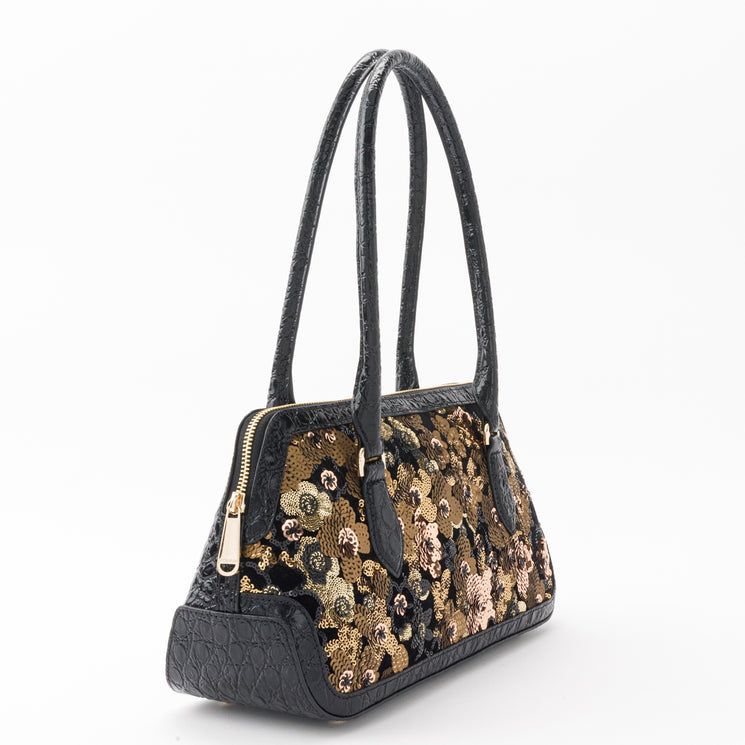 BRAHMIN Jennifer satchel with long black handles, black textured side panels, and a gold-bronze sequin floral pattern on the front. Structured silhouette shown against a plain white background.