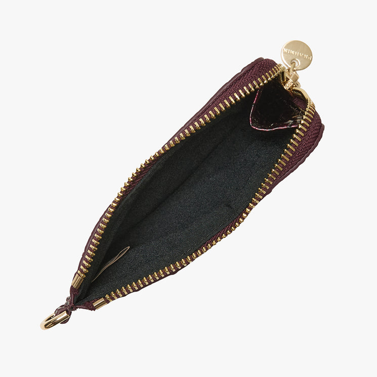 A small zippered pouch shown open against a light gray background, featuring a dark burgundy exterior, a black interior lining, and gold-tone hardware including a zipper, pull tab, and round brand tag.