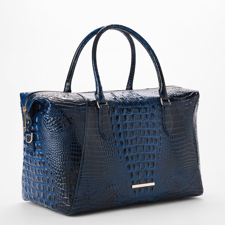 A structured navy blue crocodile-embossed leather handbag with dual top handles, gold-tone hardware, and a small horizontal gold Brahmin plaque on the front, photographed on a clean white studio background.