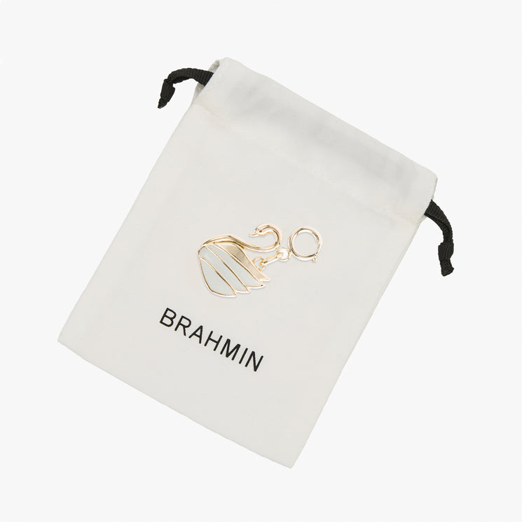 Small gold-tone swan charm with white inlay, resting on a white fabric Brahmin drawstring pouch with a black logo.