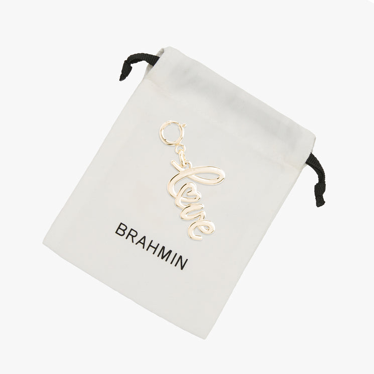 Gold-toned Love Letters script charm resting on a white fabric drawstring pouch featuring the black BRAHMIN logo.