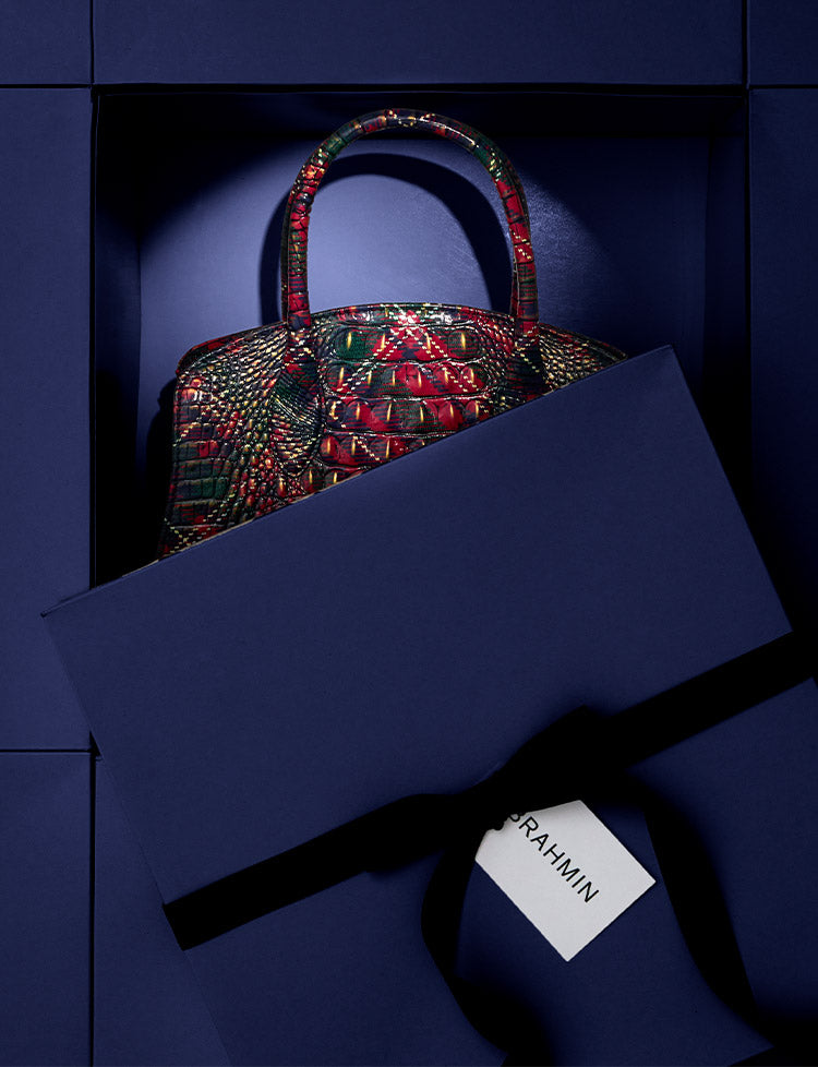 Limited-edition Brahmin satchel in a red and green croc-embossed leather, partially revealed inside a deep navy gift box with dramatic spotlighting for a holiday unboxing moment.