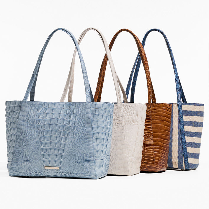 Four tall open-top totes on a white background: a light-blue crocodile-embossed leather tote in front, followed by an ivory croc-embossed tote, a cognac-brown croc-embossed tote, and a navy-and-cream striped canvas tote with blue textured trim and shoulder straps.