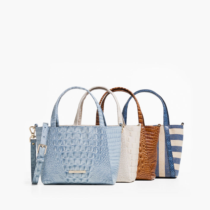 Four smaller tote bags arranged in a row on white: a light-blue crocodile-embossed mini tote with top handles and a detachable crossbody strap, an ivory croc-embossed mini tote, a cognac-brown croc-embossed mini tote, and a navy-and-cream striped canvas mini tote with blue trim and handles.”